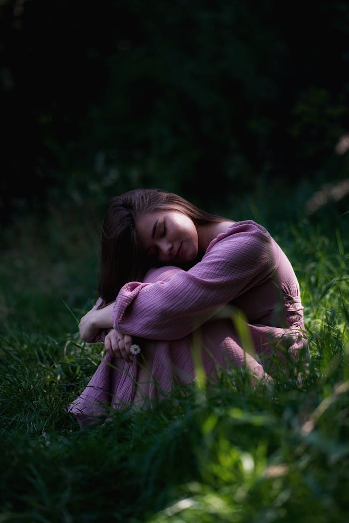 services-02 A young woman in a pink dress sits peacefully in a sunlit grassy area, embodying serenity and relaxation.