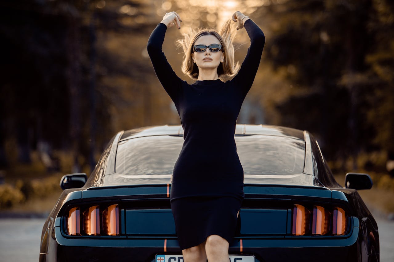 portfolio-02 Stylish woman wearing sunglasses poses confidently next to a Ford Mustang in an outdoor setting, embodying elegance and strength.